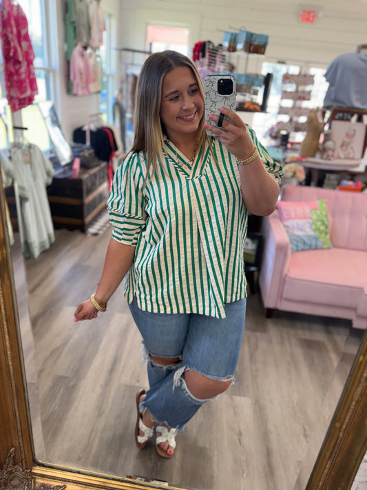 Woman taking a mirror selfie wearing a green and white striped shirt and ripped jeans in a store.