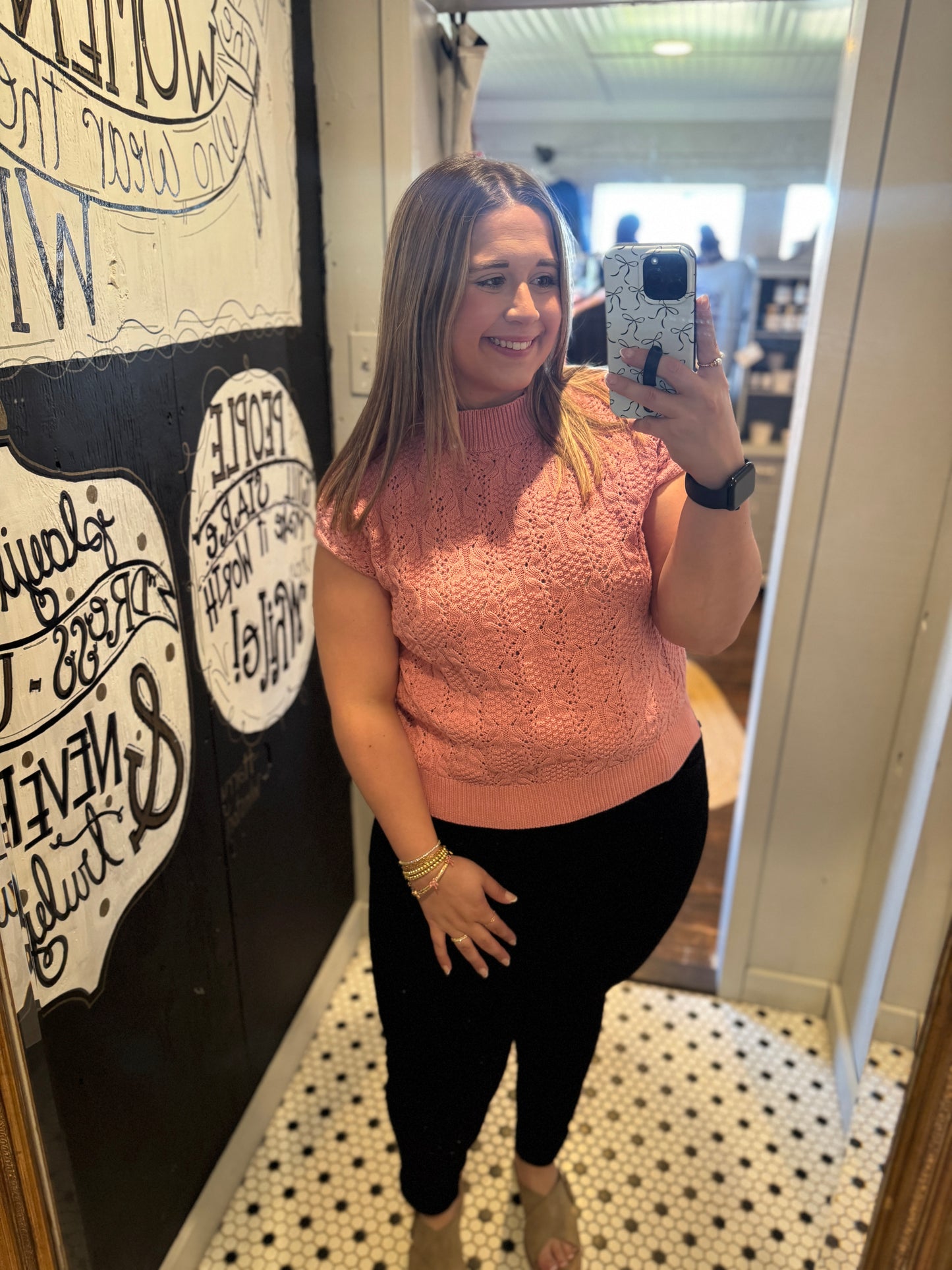 Woman taking a mirror selfie wearing a pink sleeveless top and black pants in a store.