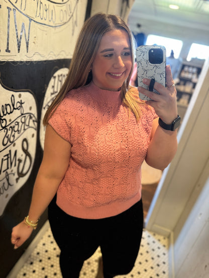 Woman taking a mirror selfie wearing a pink knitted top and black pants.