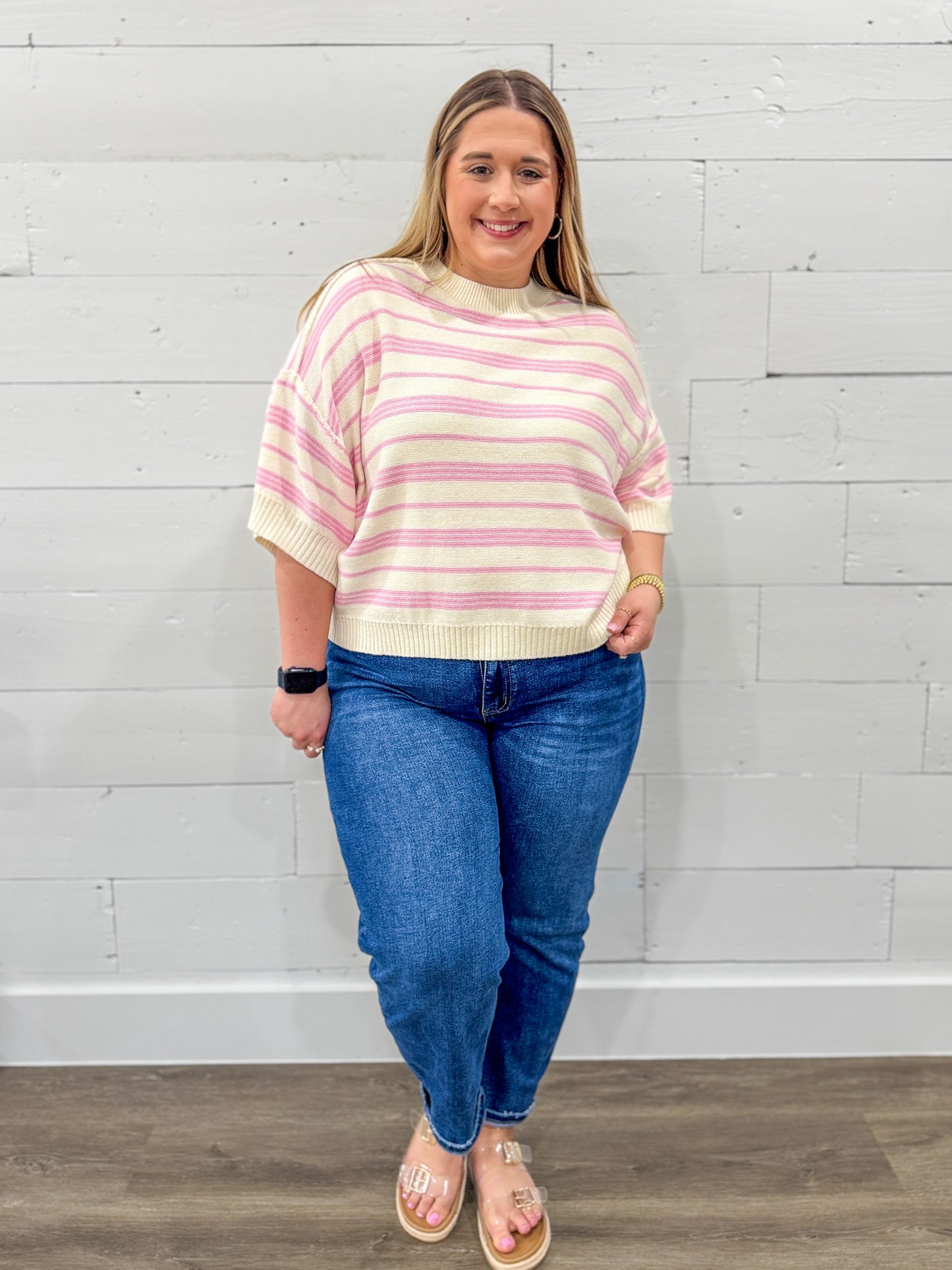Woman wearing a striped sweater and blue jeans against a white brick wall.