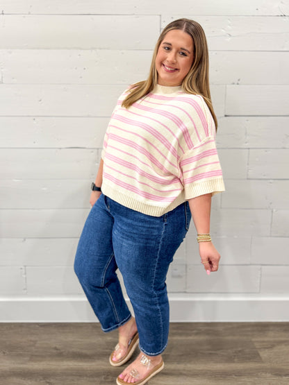 Woman wearing a pink and white striped sweater and blue jeans against a white brick wall.