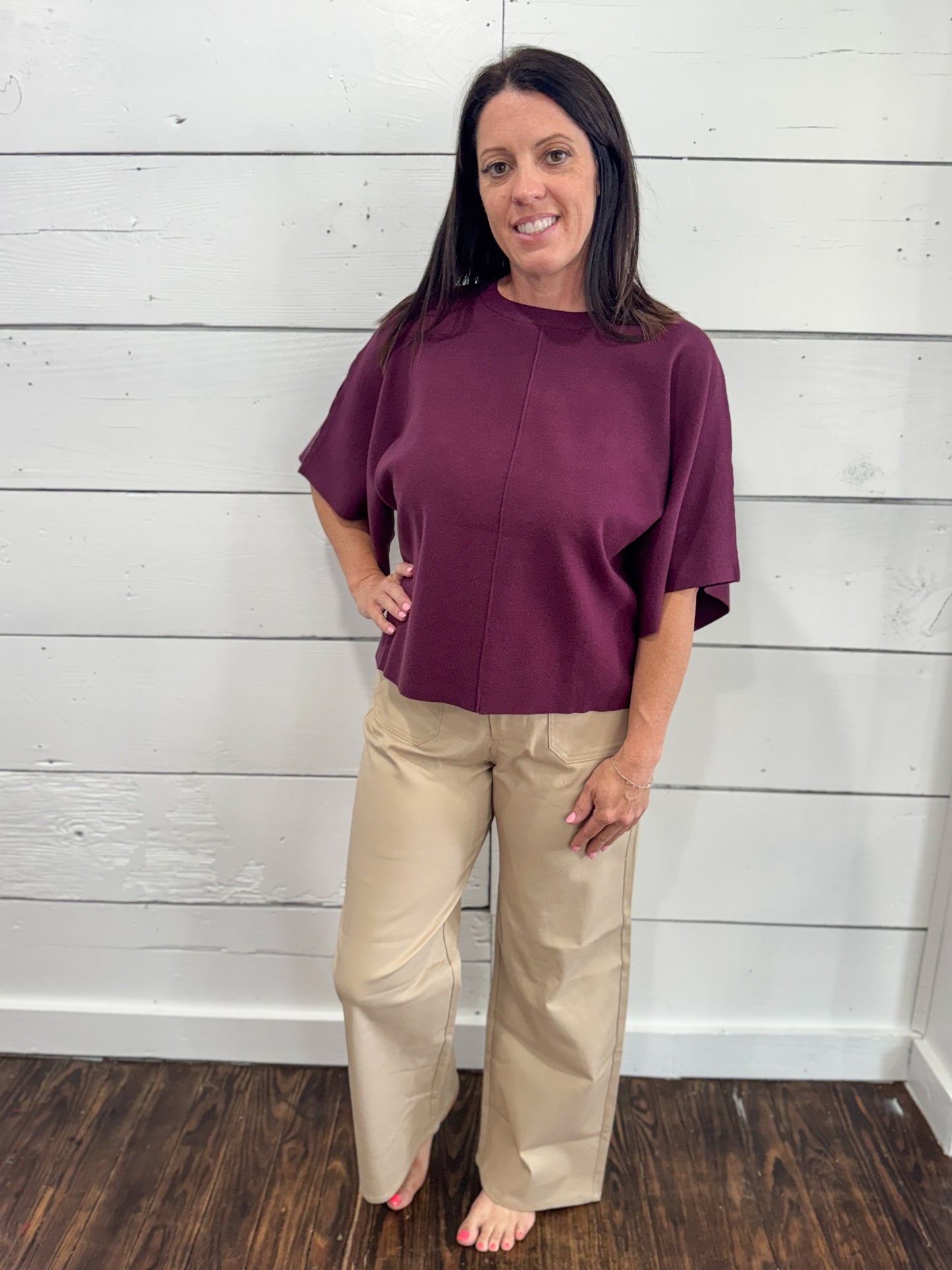 A woman stands barefoot against a white shiplap wall, wearing a short-sleeve, high-neck, front-zip mauve top and beige wide-leg pants.