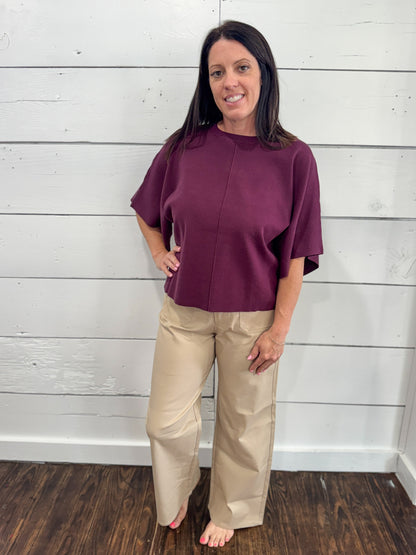 A woman stands barefoot against a white shiplap wall, wearing a short-sleeve, high-neck, front-zip mauve top and beige wide-leg pants.