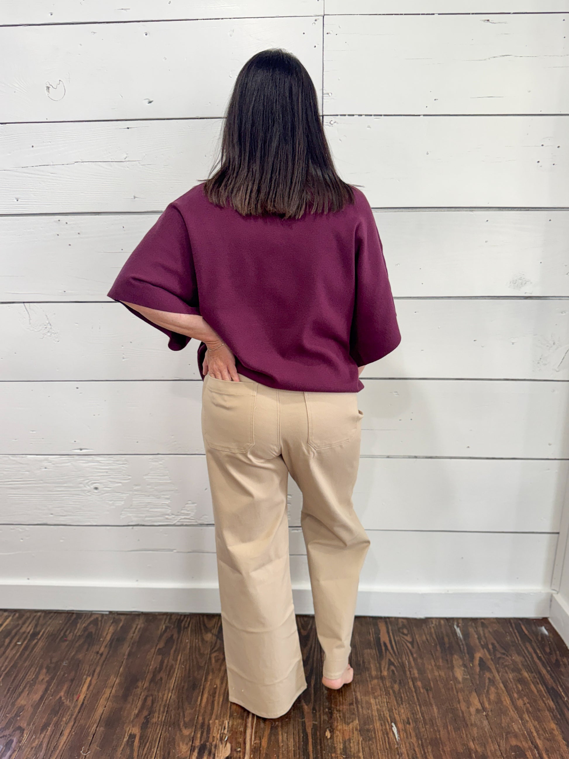 A woman stands with her back to the camera wearing a loose, short-sleeve burgundy top and beige wide-leg pants, standing on a dark wood floor against a white wood-paneled wall.