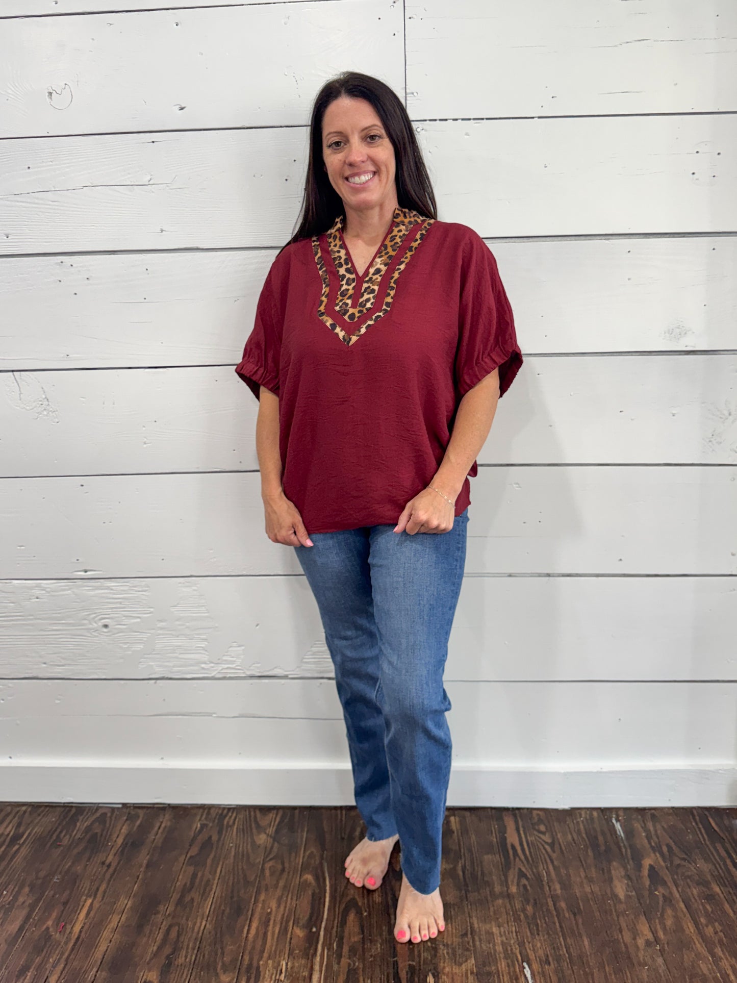 A woman modeling a short-sleeve maroon top with a decorative gold and green embroidered V-neckline, paired with blue jeans, standing against a white wood-paneled wall.