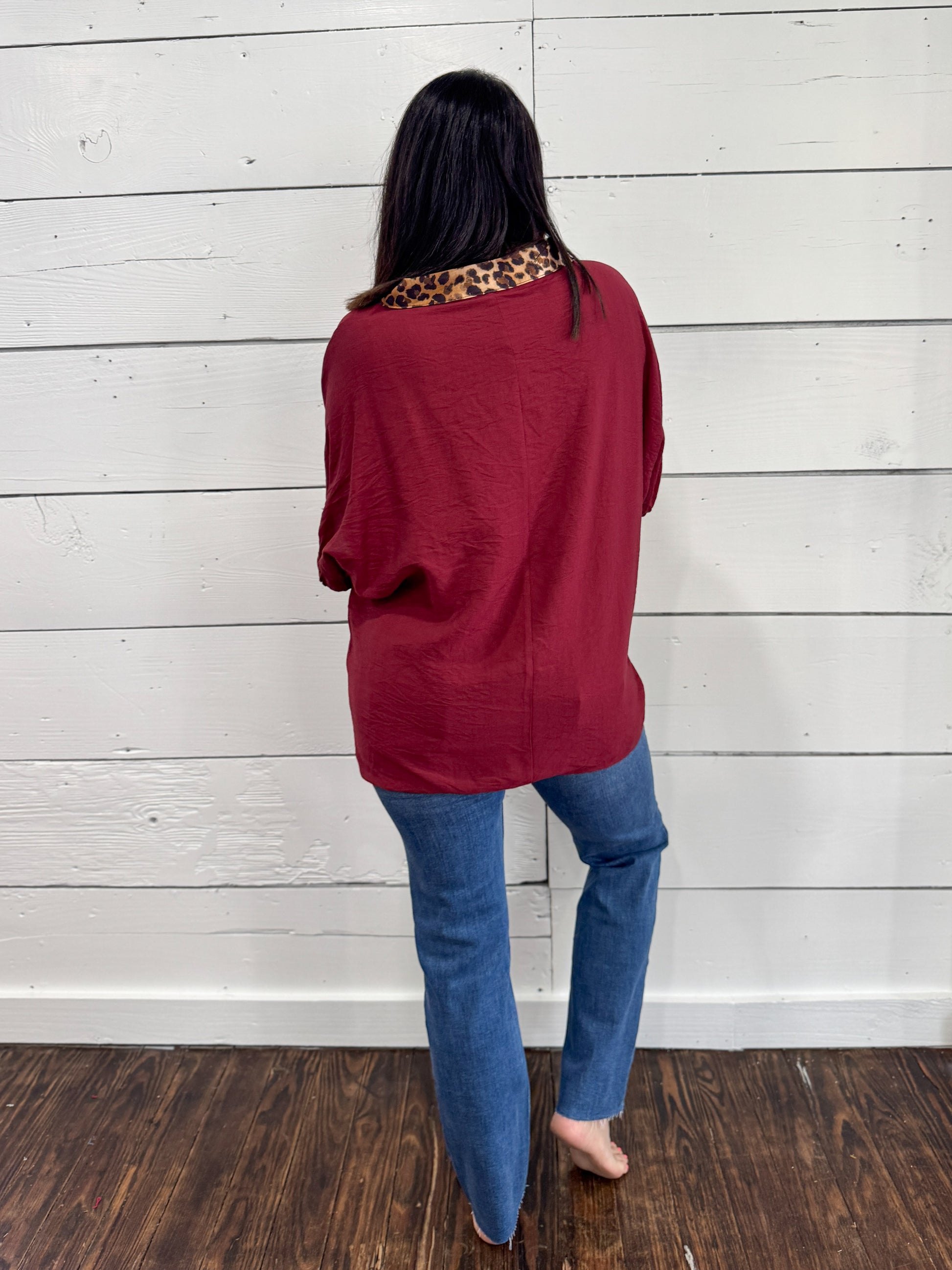 A woman viewed from behind wearing a loose-fitting burgundy top with a leopard-print collar, paired with blue jeans, standing against a white wood-paneled wall.