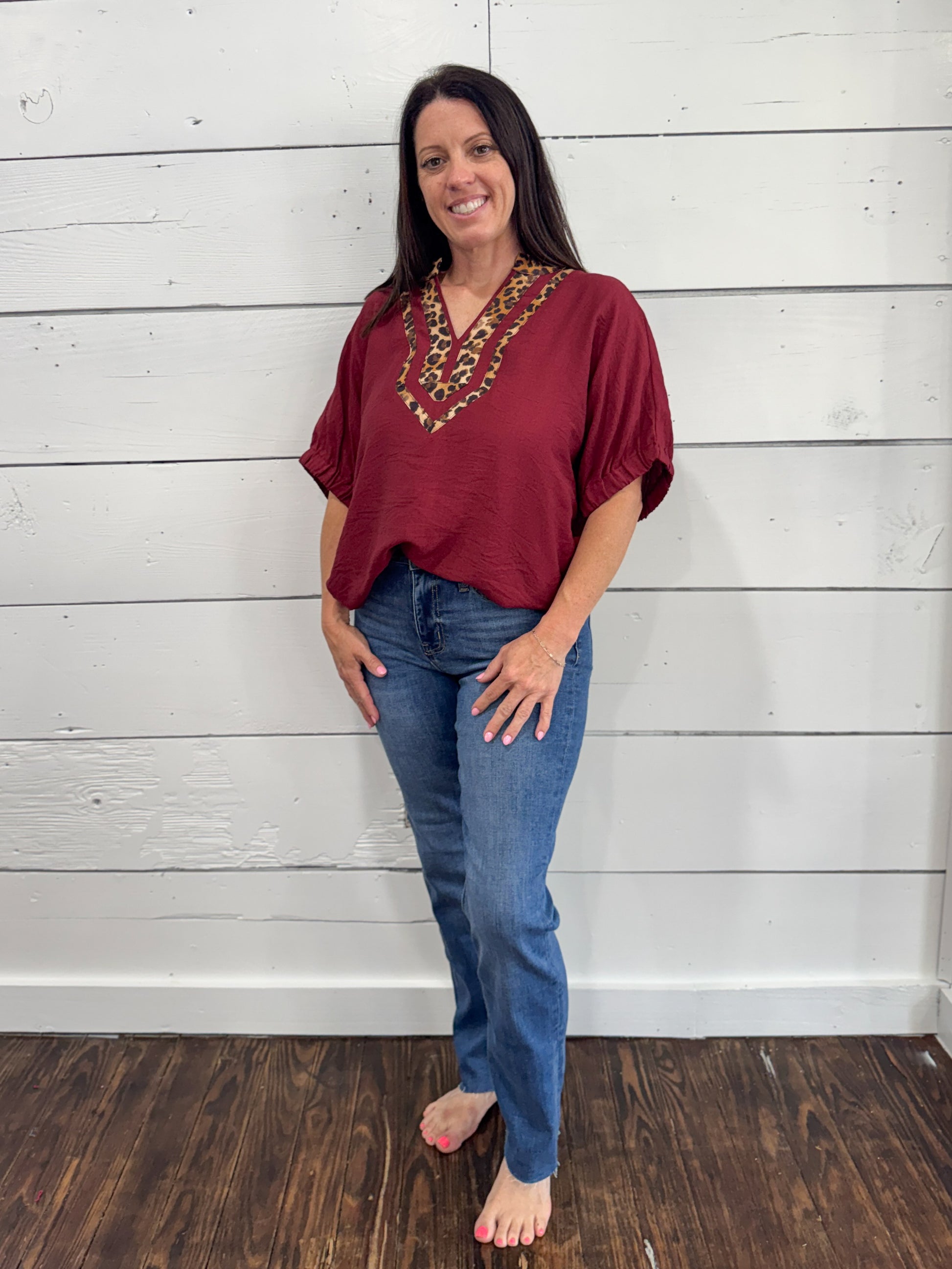 A woman modeling a short-sleeve maroon top with a decorative embroidered yoke in red, gold, and green, paired with blue jeans, standing against a white wood-paneled wall.