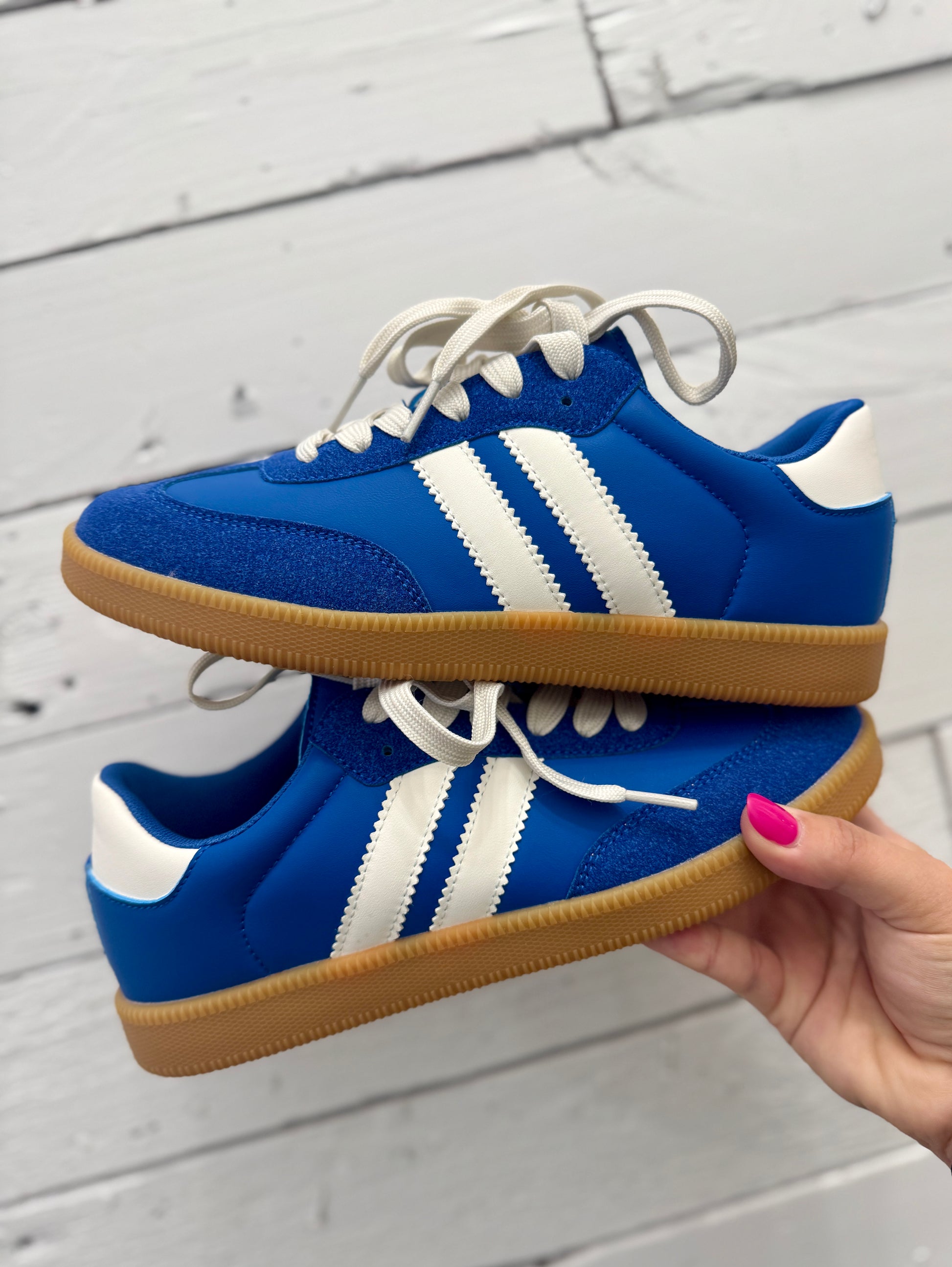 A pair of blue low-top sneakers with white side stripes, white laces, and gum soles, held by a hand against a white wood plank background.