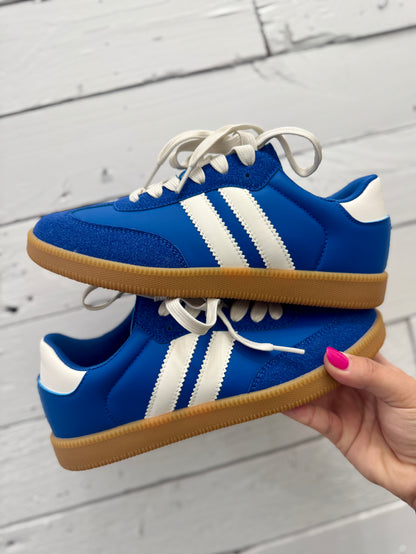 A pair of blue low-top sneakers with white side stripes, white laces, and gum soles, held by a hand against a white wood plank background.
