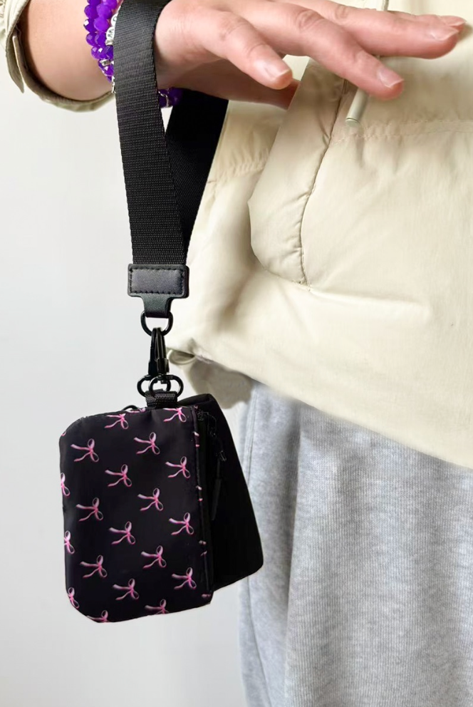 Black pouch with pink patterns attached to a strap, held by a person wearing a beige jacket.