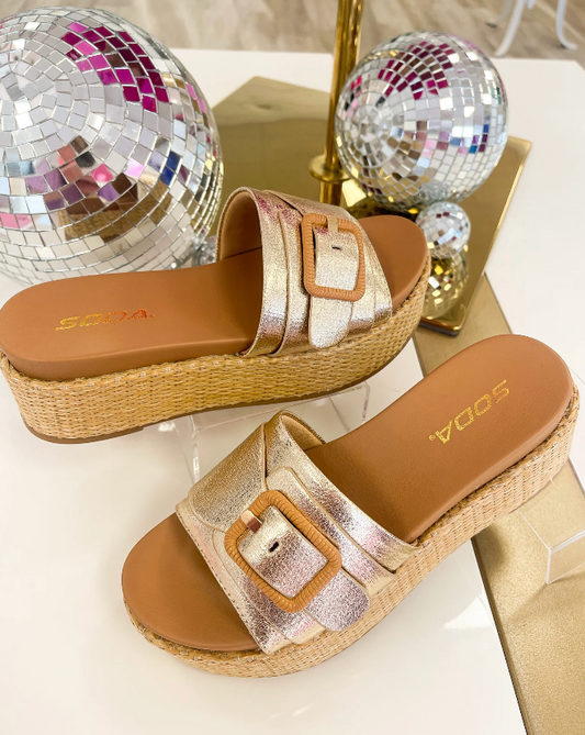 Gold sandals with woven sole and brown insole on a white surface with disco balls in the background.
