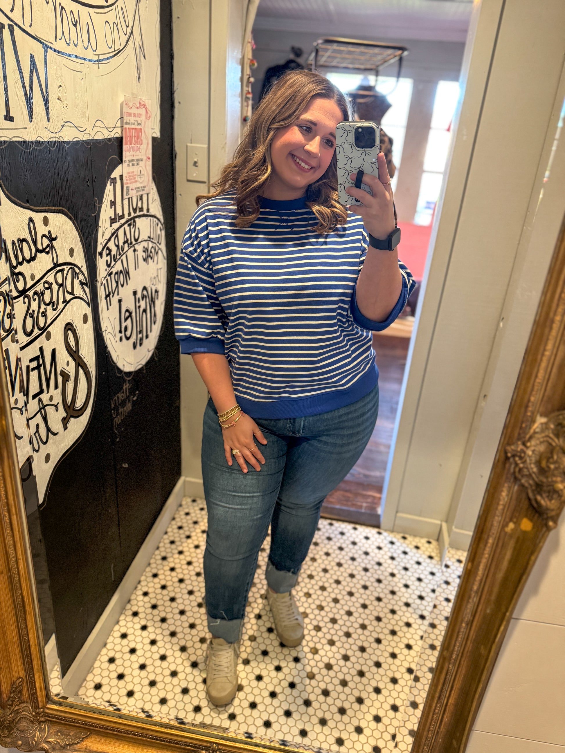 Woman taking a mirror selfie wearing a blue and white striped shirt and jeans in a room with a decorative wall.