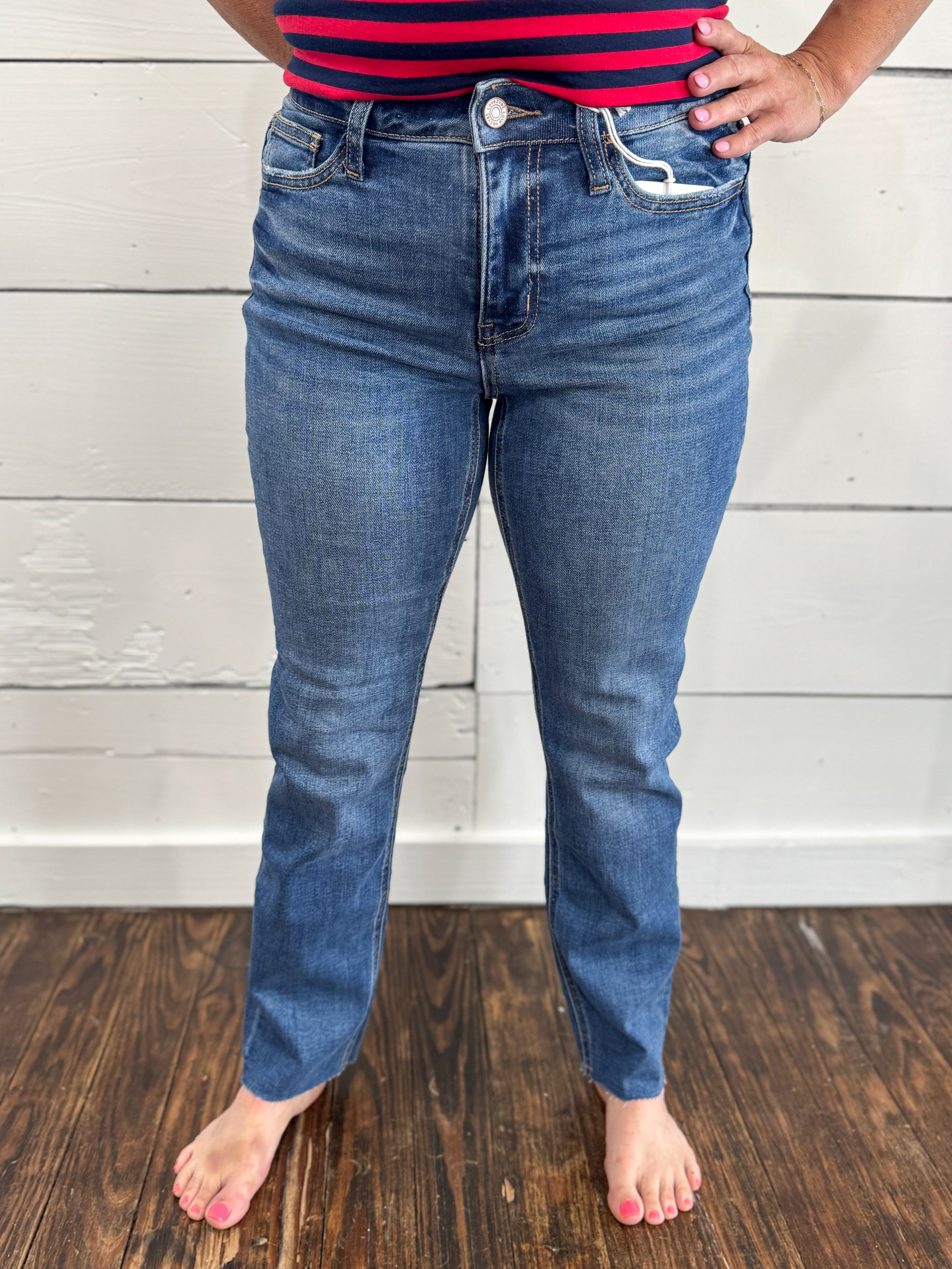 A person wearing medium-wash blue jeans, shown from the waist down, standing barefoot on a dark wood floor against a white wood-paneled wall.
