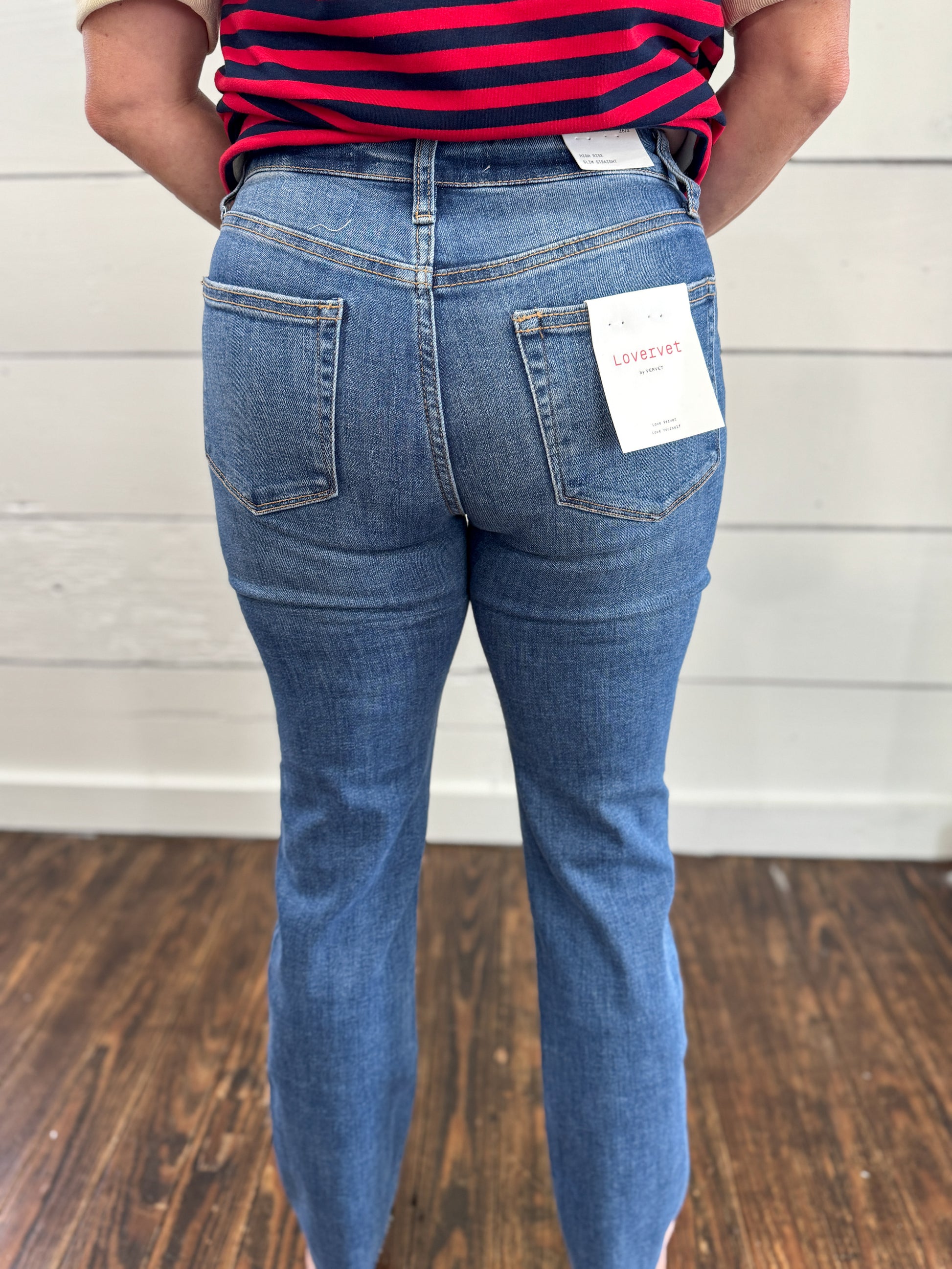 A person shown from the back wearing medium-wash blue jeans with a white Lovezite brand tag attached to the back pocket, standing on a wood floor against a white paneled wall.
