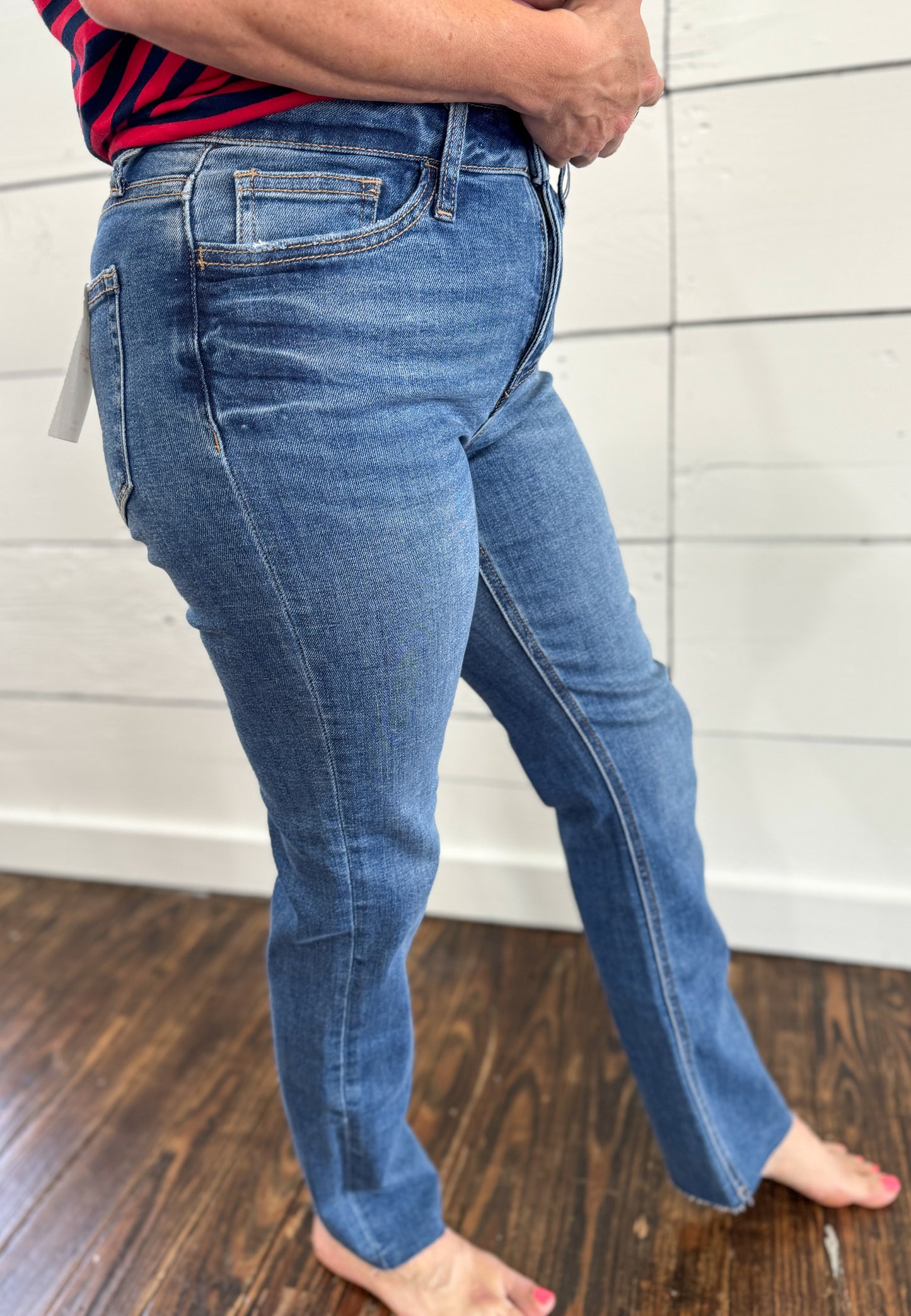 A person wearing medium-wash blue denim jeans, shown from the waist down, standing barefoot on a dark wood floor against a white paneled wall.