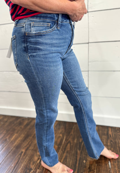 A person wearing medium-wash blue denim jeans, shown from the waist down, standing barefoot on a dark wood floor against a white paneled wall.