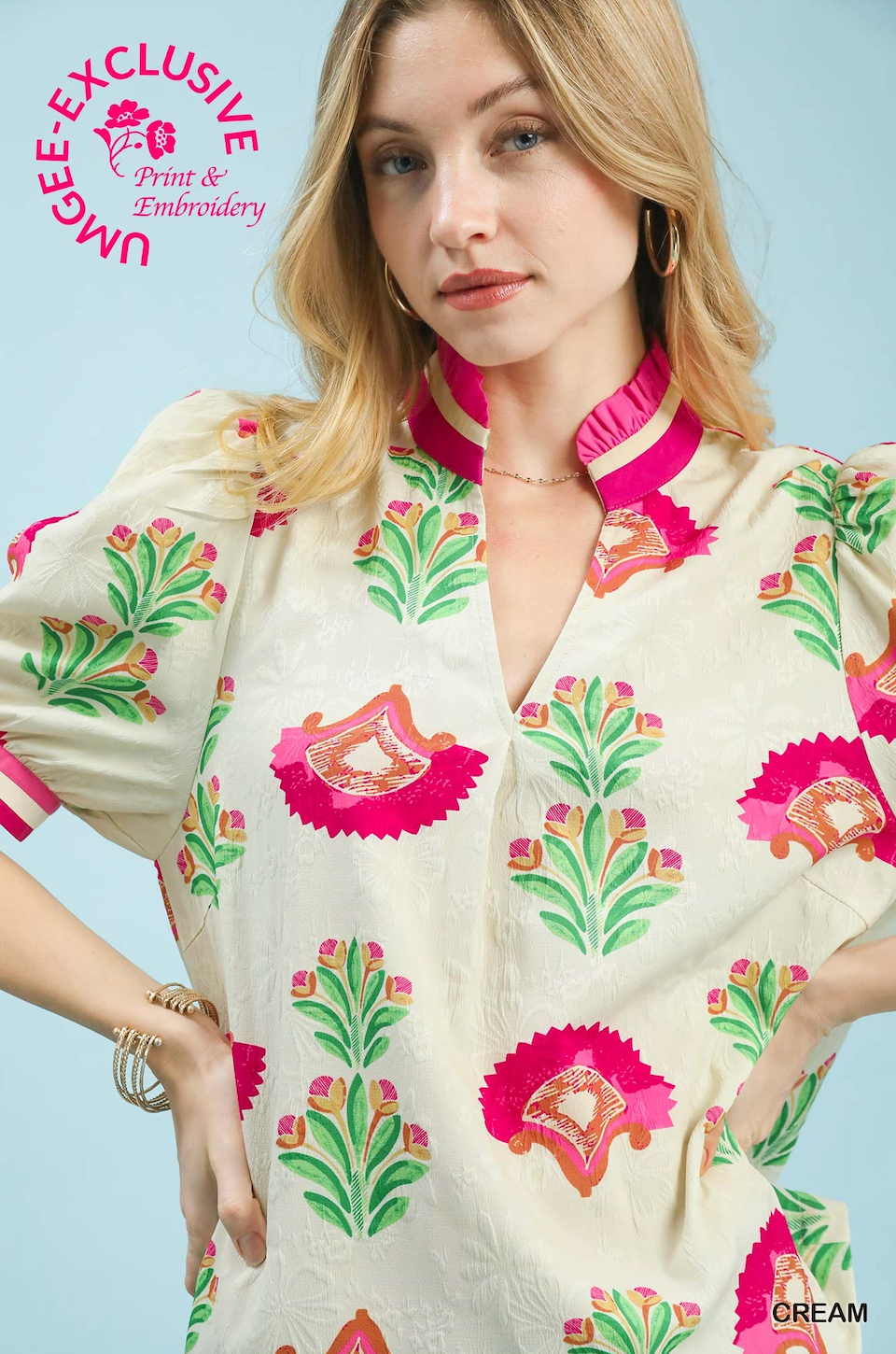 Woman wearing a cream-colored blouse with pink and green floral embroidery against a light blue background.