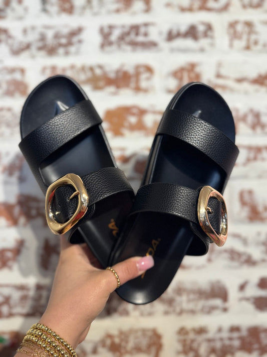 Black sandals with gold buckles held by a hand against a textured wall background