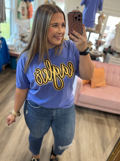 A woman taking a mirror selfie in a boutique, wearing a blue crewneck t-shirt with a gold and black script graphic that reads 'savage'.