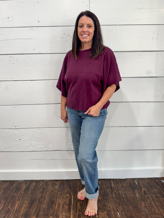 A woman stands barefoot against a white wood plank wall, wearing a loose short-sleeve crewneck top in deep burgundy and light blue jeans.