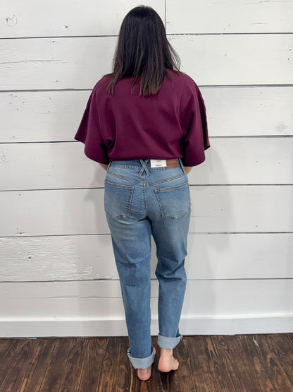 A person viewed from behind wearing a dark burgundy short-sleeve crop top and light blue jeans, standing barefoot on a dark wood floor against a white wood-paneled wall.