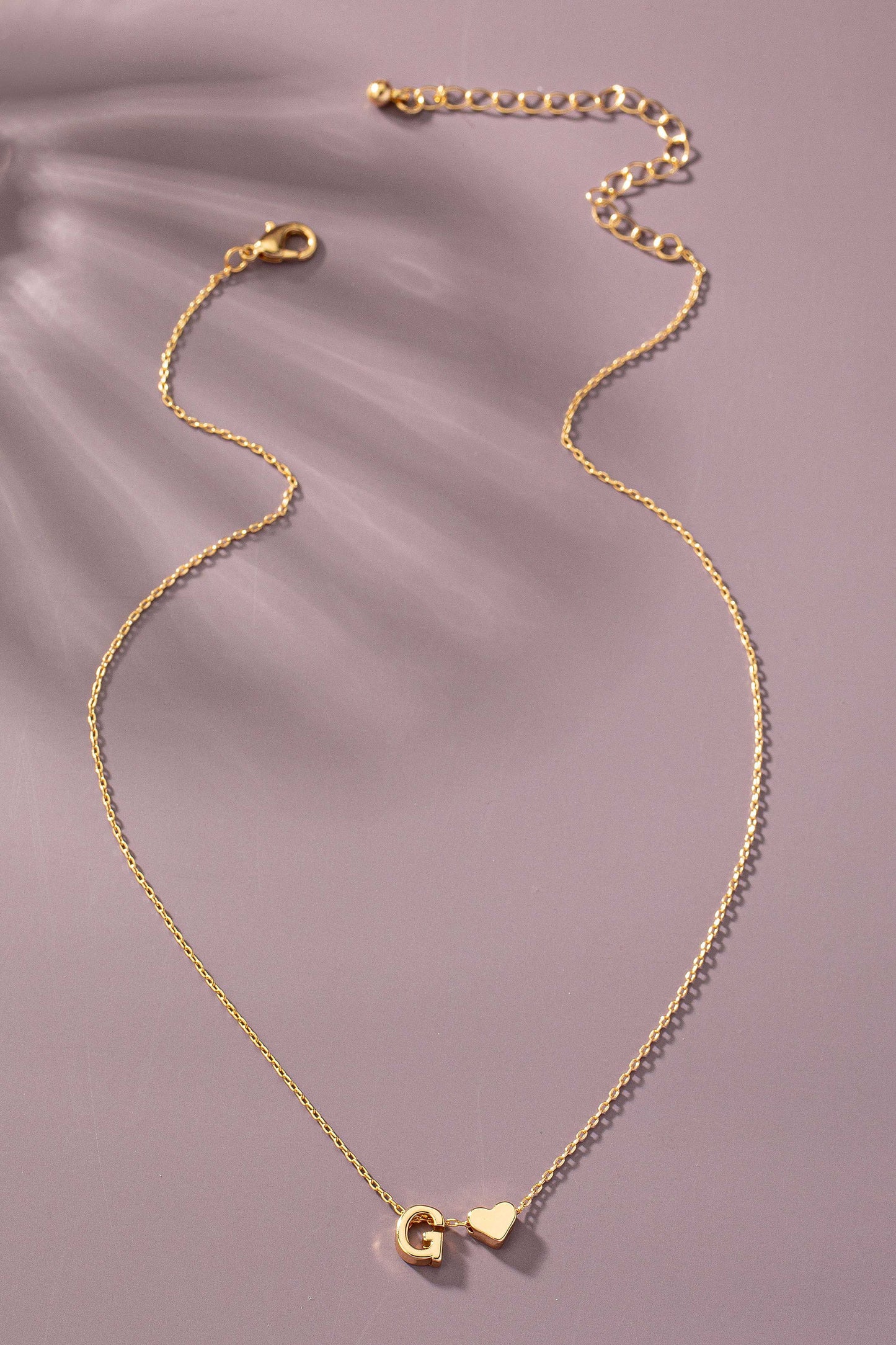 Gold necklace with 'G' and heart pendant on a soft purple background