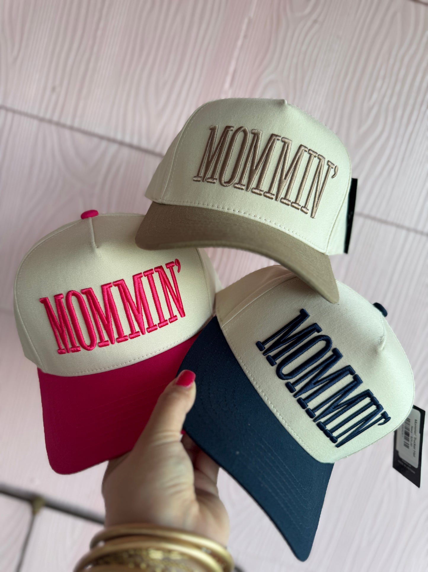 Three caps with 'MOMMY' and 'MOMMIN' text held by a hand on a light wooden surface.