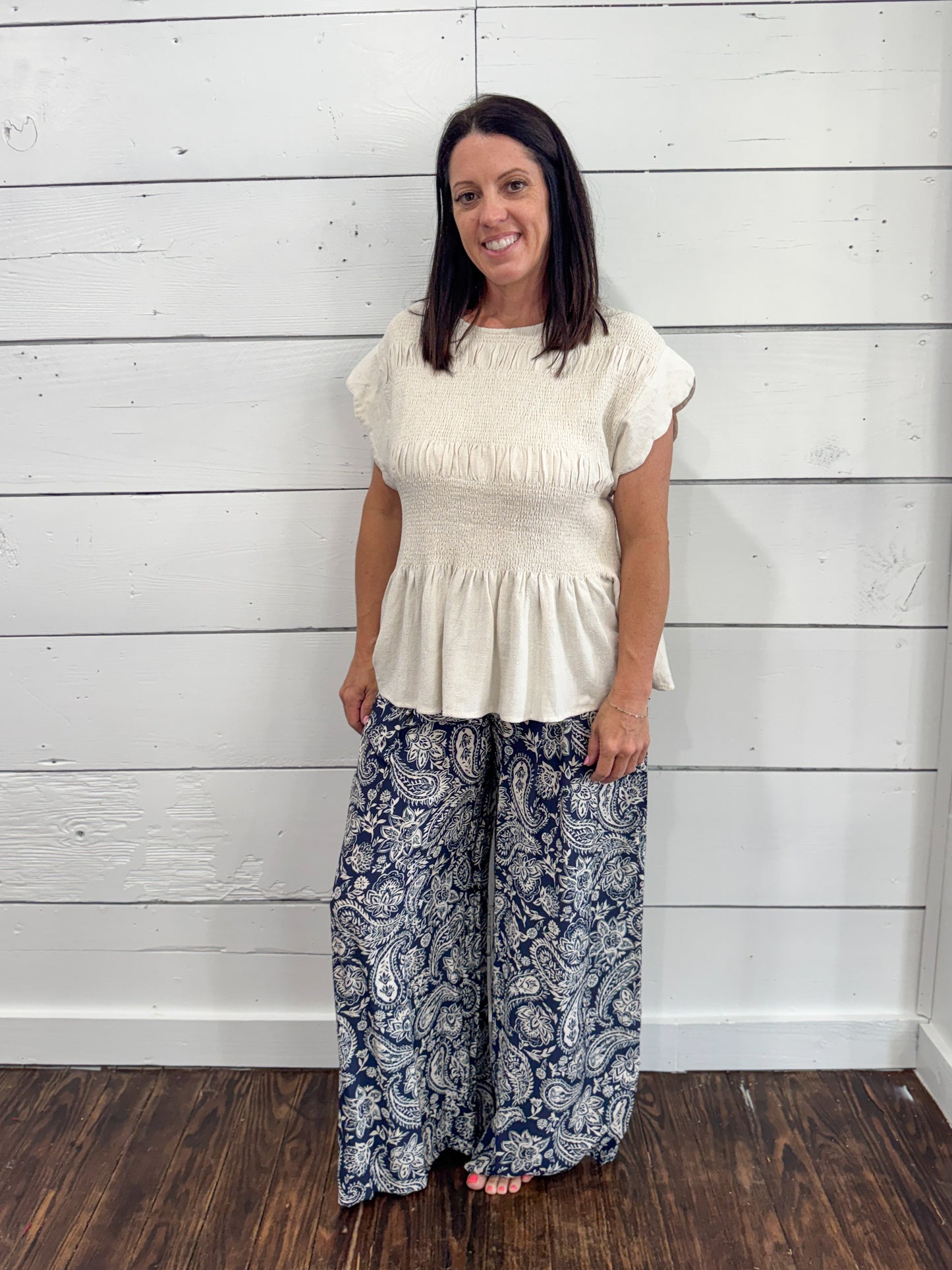 A woman modeling a cream-colored short-sleeve peplum top paired with navy blue and white paisley wide-leg pants, standing against a white wood-paneled wall.
