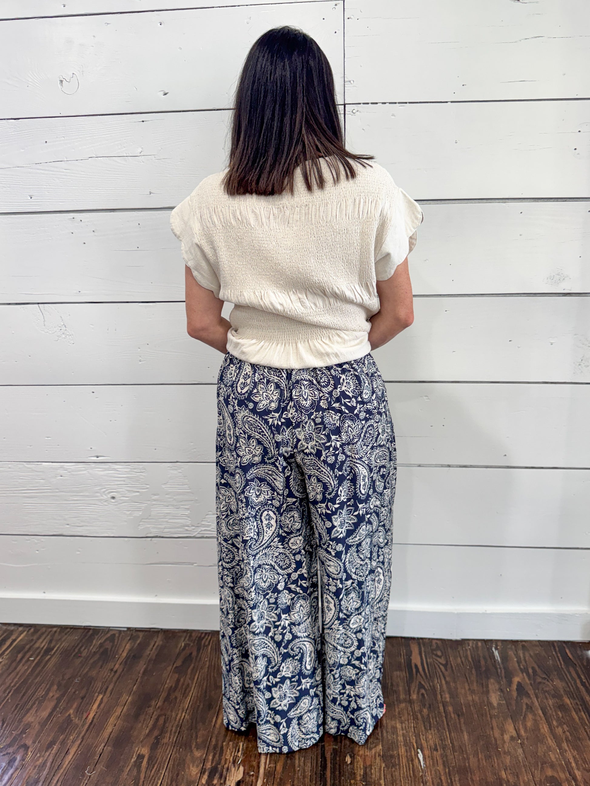 A woman viewed from behind wearing a cream-colored textured short-sleeve top and navy blue wide-leg pants with a white paisley pattern, standing against a white wood-paneled wall on a dark wood floor.