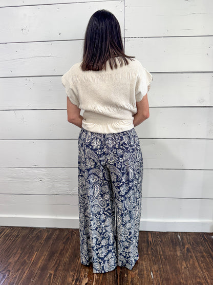 A woman viewed from behind wearing a cream-colored textured short-sleeve top and navy blue wide-leg pants with a white paisley pattern, standing against a white wood-paneled wall on a dark wood floor.