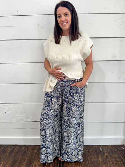 A woman modeling a cream-colored short-sleeve fringed top and navy blue wide-leg pants with a white paisley pattern, standing against a white wood-paneled wall.
