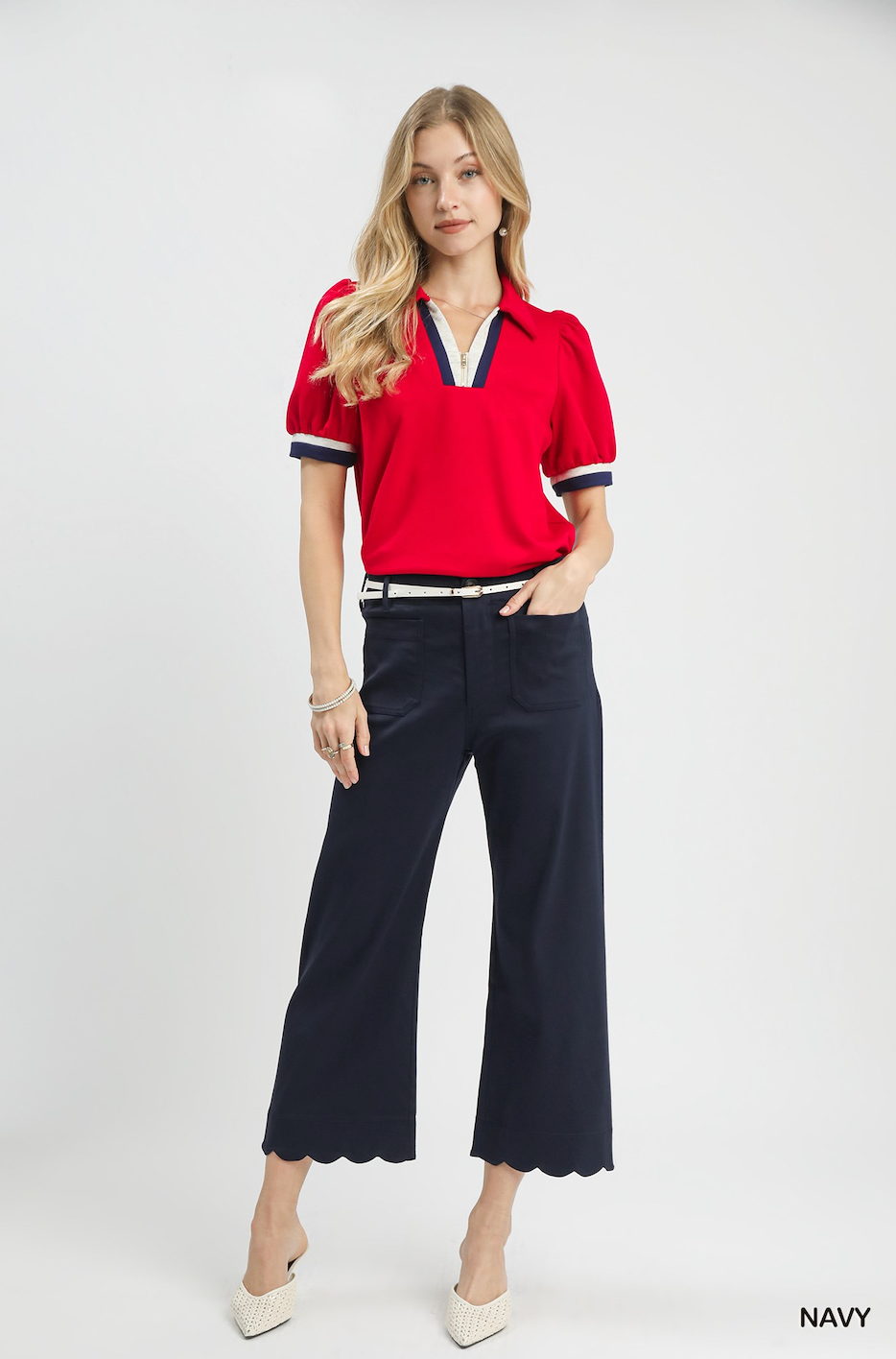 A woman modeling a red short-sleeve polo shirt with navy and white trim, paired with navy cropped pants and white heels, against a light gray studio background.