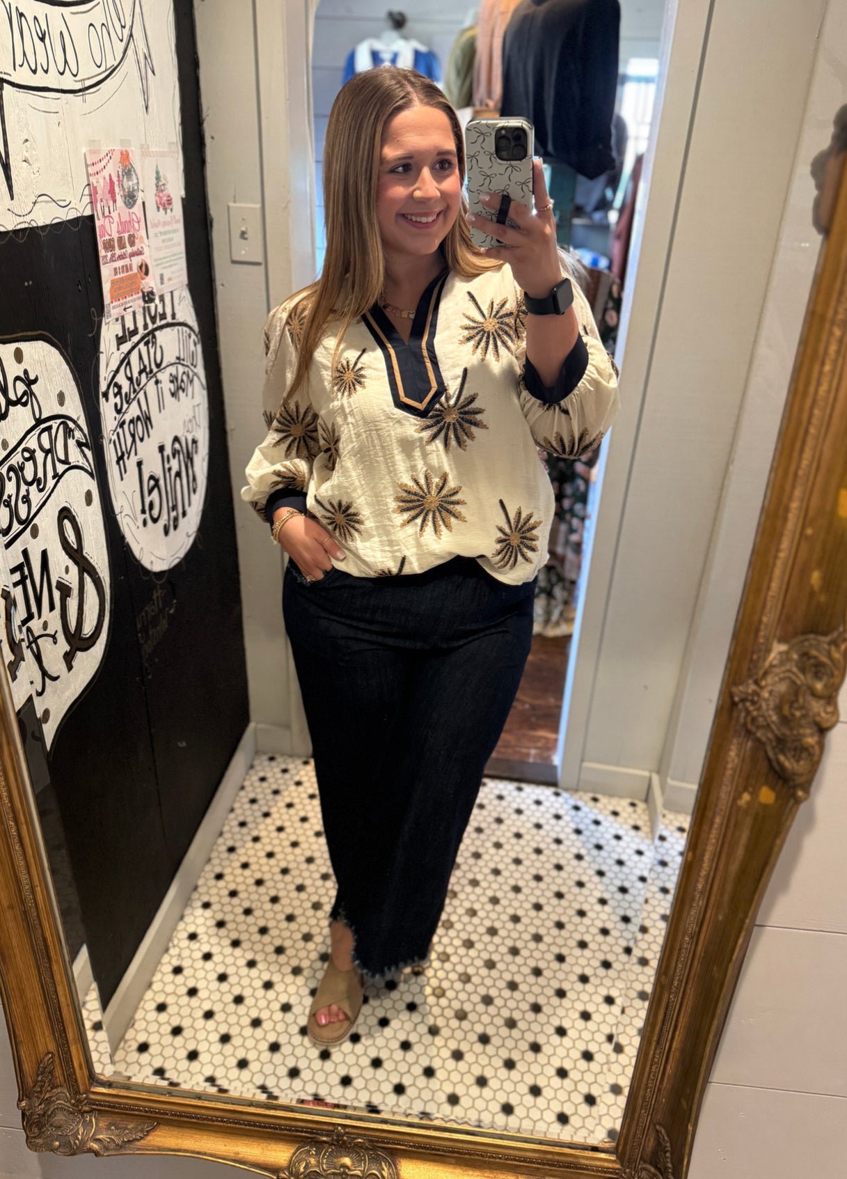 Woman taking a mirror selfie wearing a white blouse with black patterns and black pants in a room with a decorative mirror.