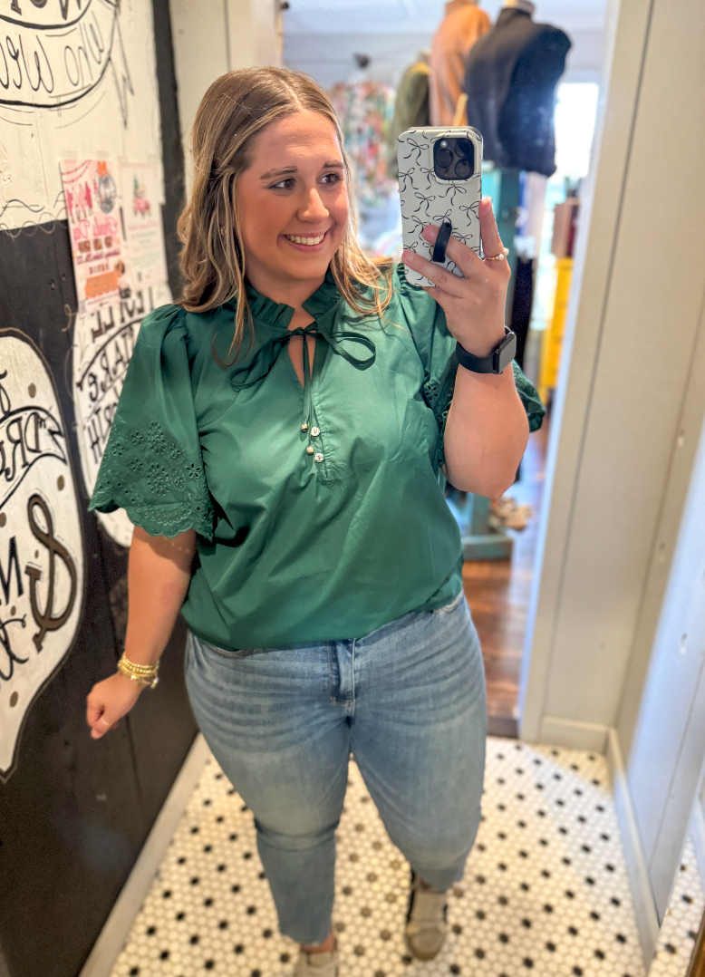 Woman taking a mirror selfie wearing a green blouse and blue jeans in a store.