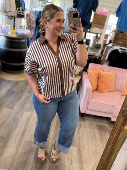 A woman takes a mirror selfie in a boutique while wearing a brown and white vertically striped short-sleeve button-up shirt with blue jeans and beige sandals.
