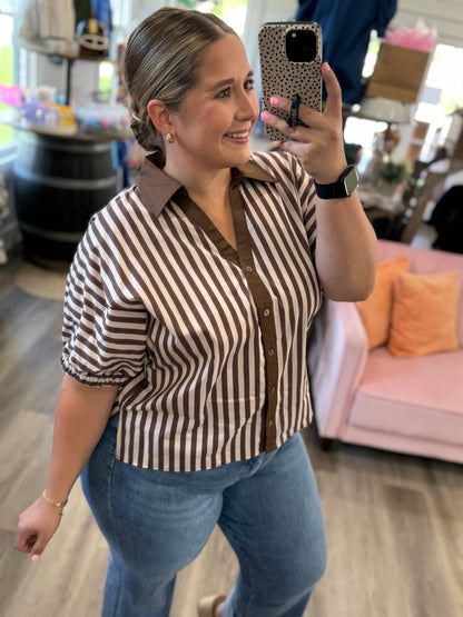 A woman takes a mirror selfie in a boutique while wearing a brown and beige vertically striped short-sleeve button-up shirt with a brown collar and blue jeans.