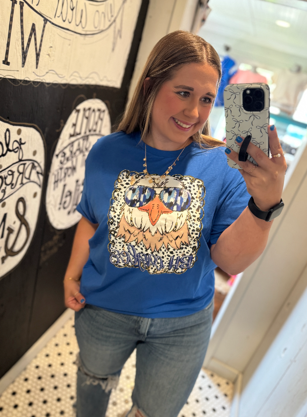 Woman taking a mirror selfie wearing a blue t-shirt with a graphic design, standing in front of a wall with text.