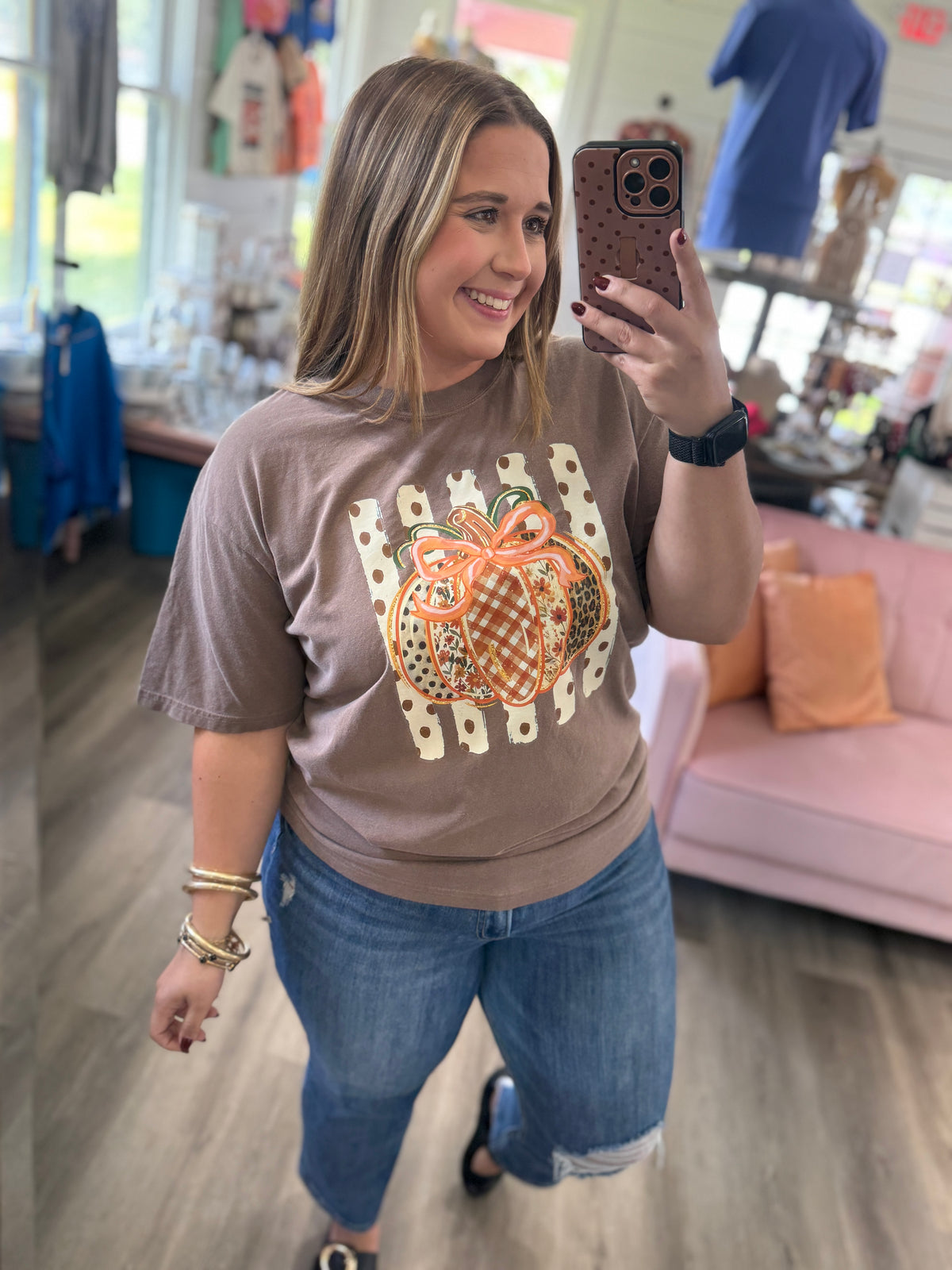 A woman taking a mirror selfie in a boutique, wearing a taupe brown t-shirt with a pumpkin graphic and blue jeans.