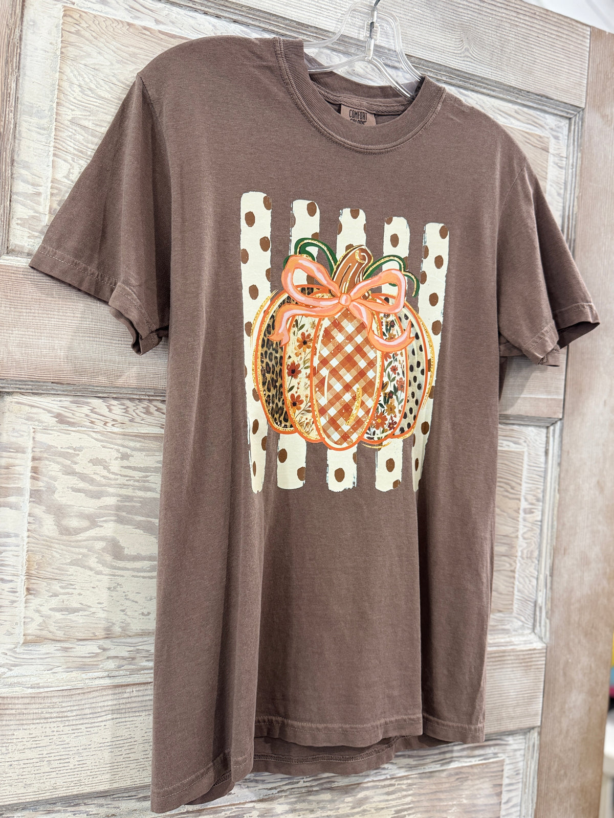 A brown short-sleeve t-shirt hanging on a hanger against a rustic wooden door. The shirt features a central graphic of a patterned orange pumpkin with a pink bow, set against a white fence motif with brown polka dots.
