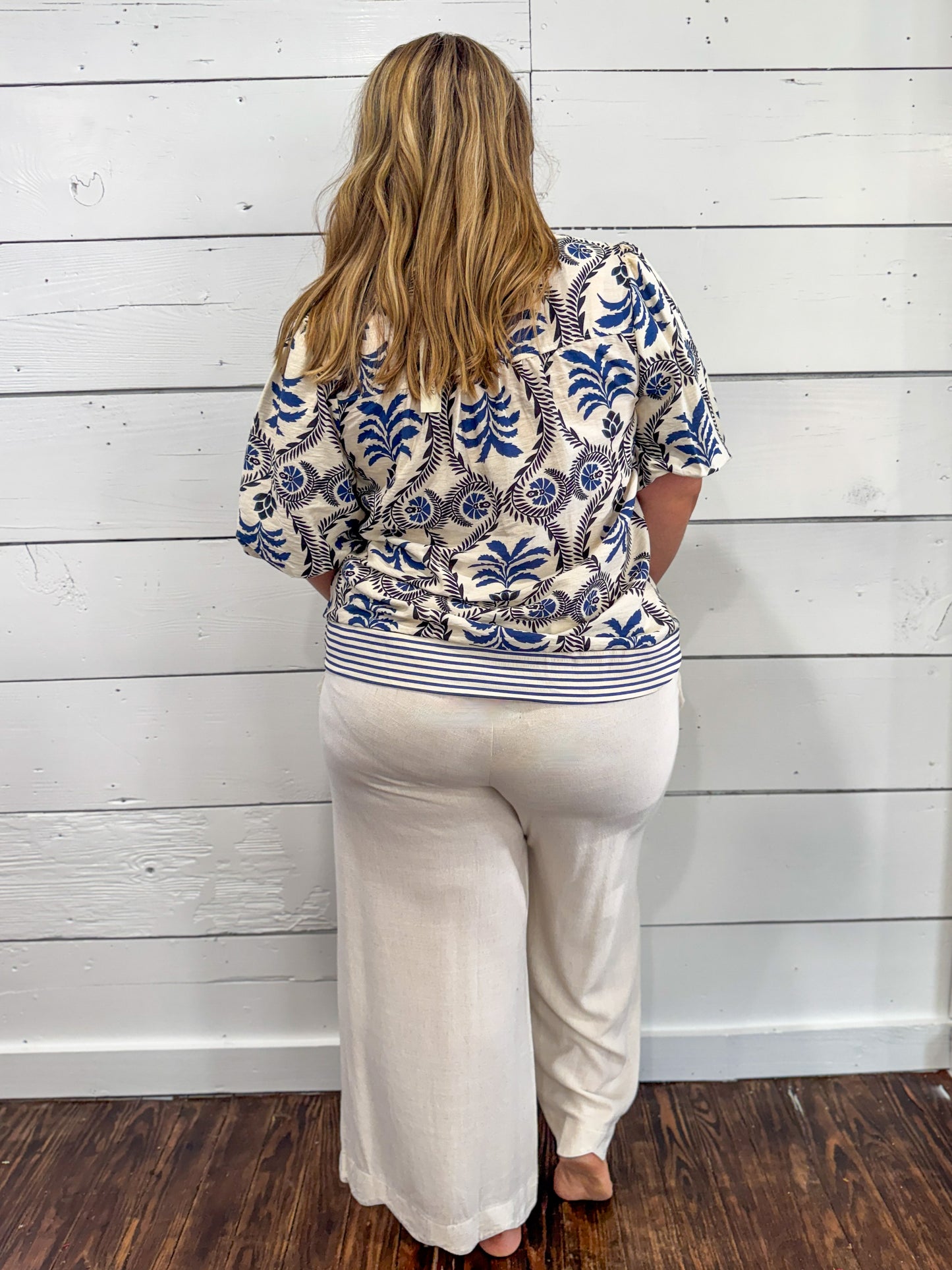 A woman viewed from behind wearing a cream-colored short-sleeve top with a blue and brown palm tree pattern and white wide-leg pants, standing against a white wood-paneled wall.