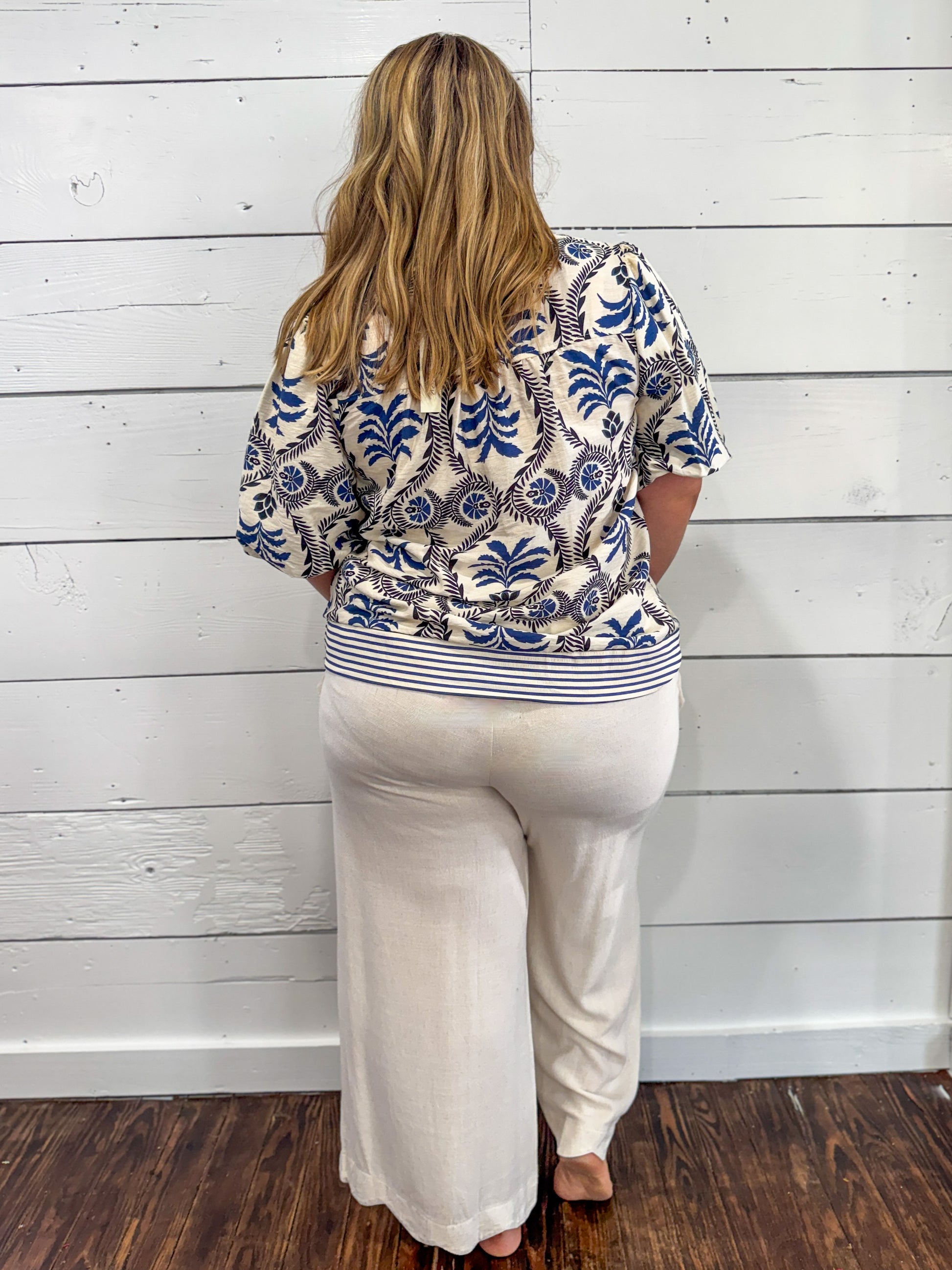 A woman viewed from behind wearing a cream-colored short-sleeve top with a blue and brown palm tree pattern and white wide-leg pants, standing against a white wood-paneled wall.