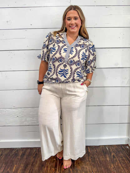 A woman modeling a short-sleeve button-up blouse with a blue and white botanical pattern on a cream background, paired with cream wide-leg pants, standing against a white wood-paneled wall.