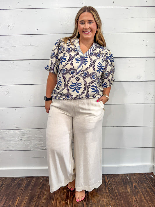 A woman modeling a short-sleeve button-up blouse with a blue and white botanical pattern on a cream background, paired with cream wide-leg pants, standing against a white wood-paneled wall.