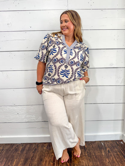 A woman modeling a short-sleeve collared blouse with a blue and white paisley pattern, paired with cream wide-leg pants, standing against a white wood-paneled wall.
