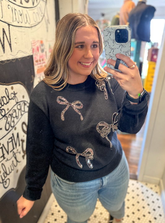 Woman taking a mirror selfie wearing a black sweater with bow designs.