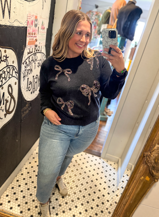 Woman taking a mirror selfie wearing a black sweater with bow patterns and blue jeans.