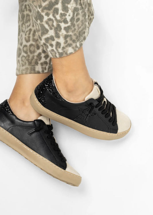 Black sneakers with beige soles worn by a person with leopard print pants on a white background