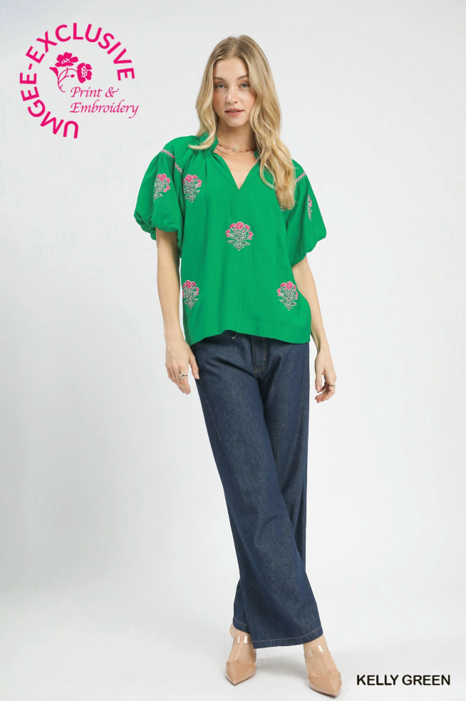 Woman wearing a green blouse with floral embroidery and blue pants on a white background.