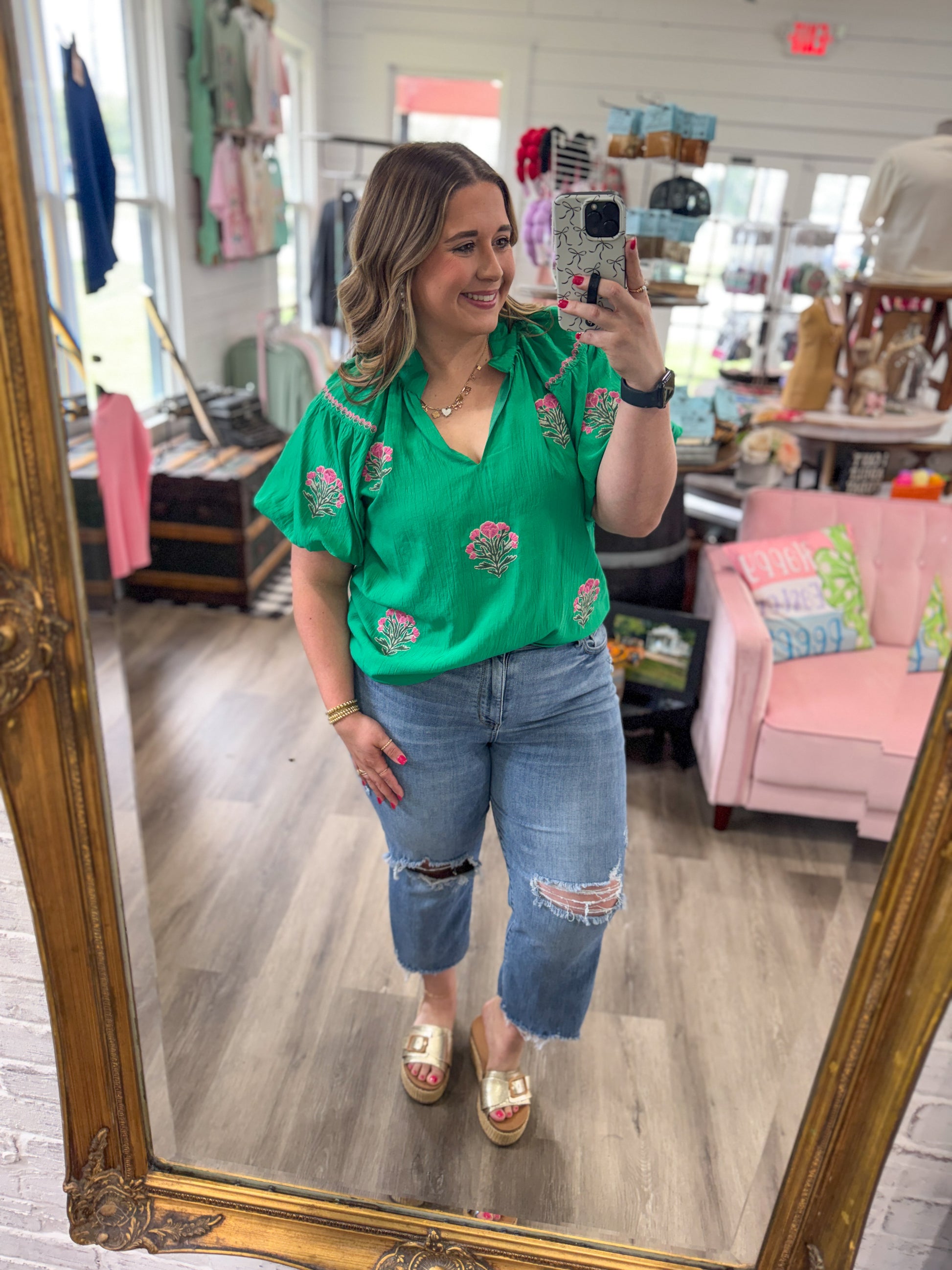 Woman taking a mirror selfie wearing a green floral blouse and blue jeans in a store.