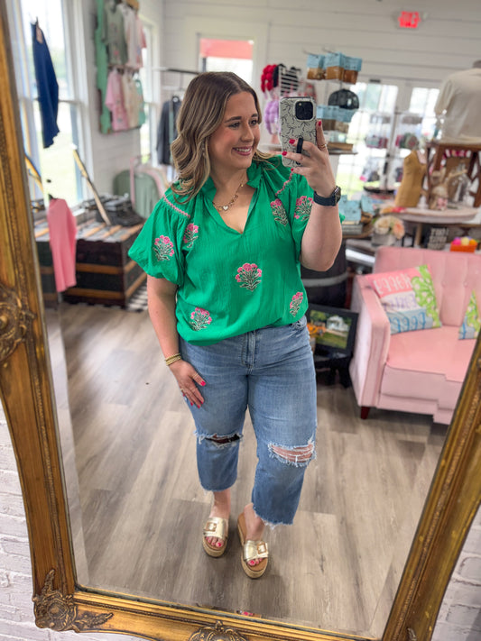 Woman taking a mirror selfie wearing a green floral blouse and blue jeans in a store.